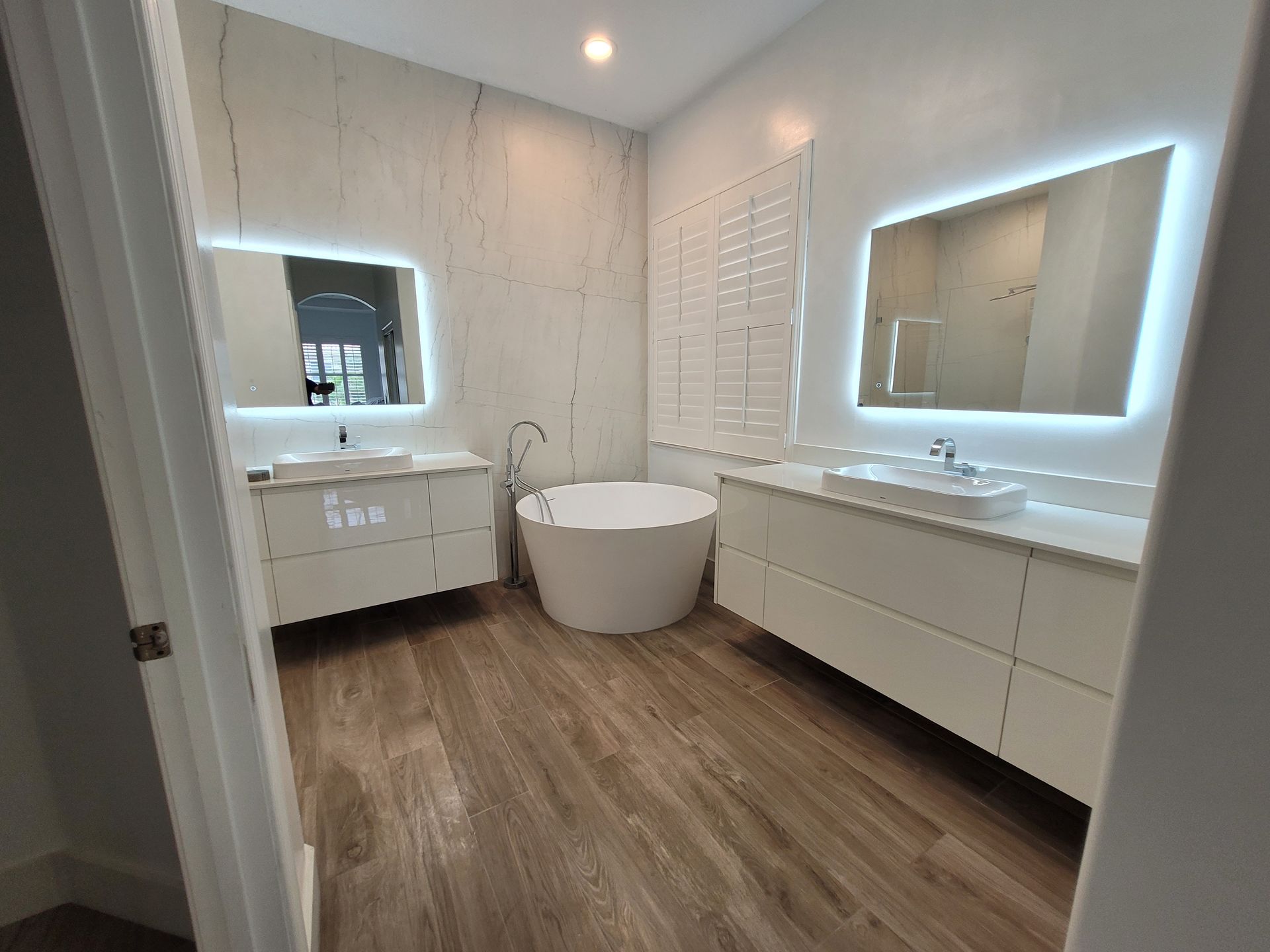 Modern bathroom with white cabinets, freestanding tub, two sinks, mirrors, and wood-look flooring.