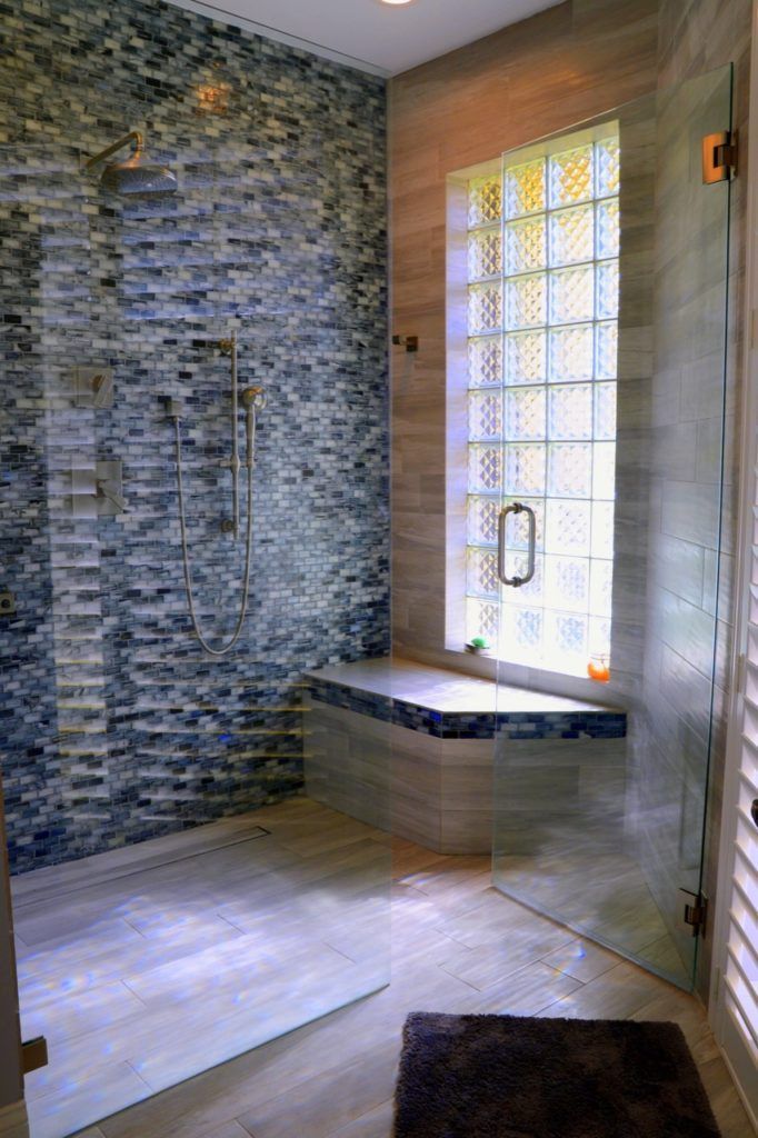 Shower with blue and grey tiled wall, glass door, window with glass blocks, and built-in bench.