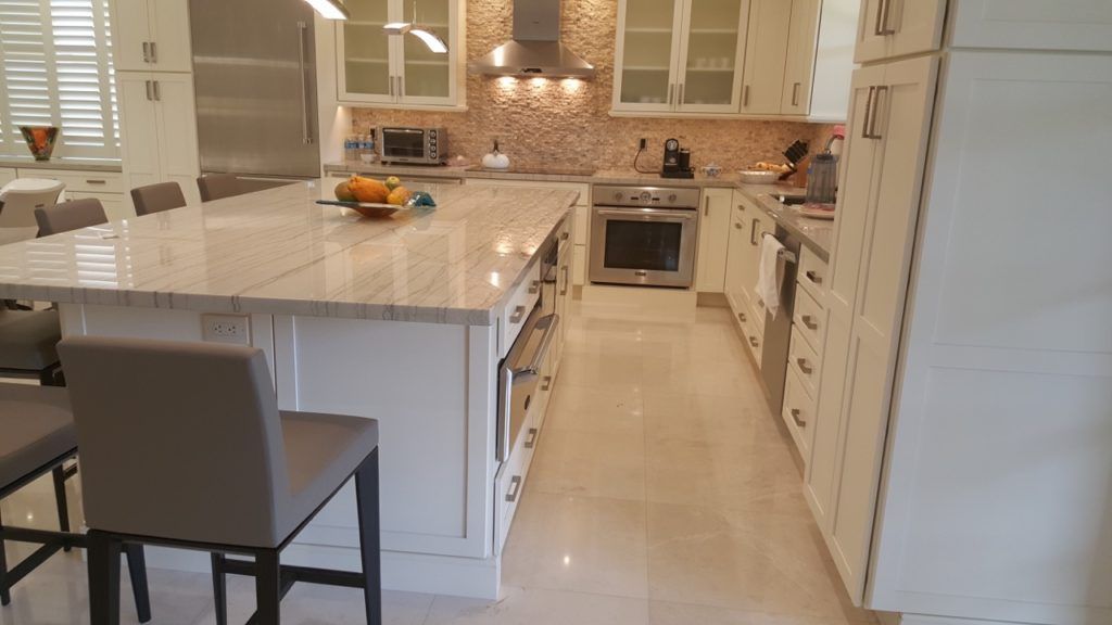 Bright kitchen with white cabinets, marble island, and beige tile floor; dining chair in foreground.