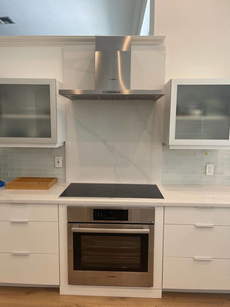 Modern kitchen with stainless steel range hood, black cooktop, and built-in oven between white cabinets