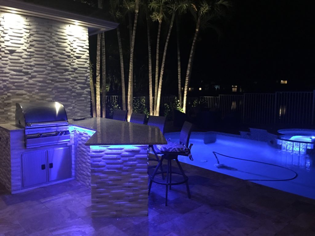 Blue-lit backyard pool and patio with grill, stone bar, and palm trees at night