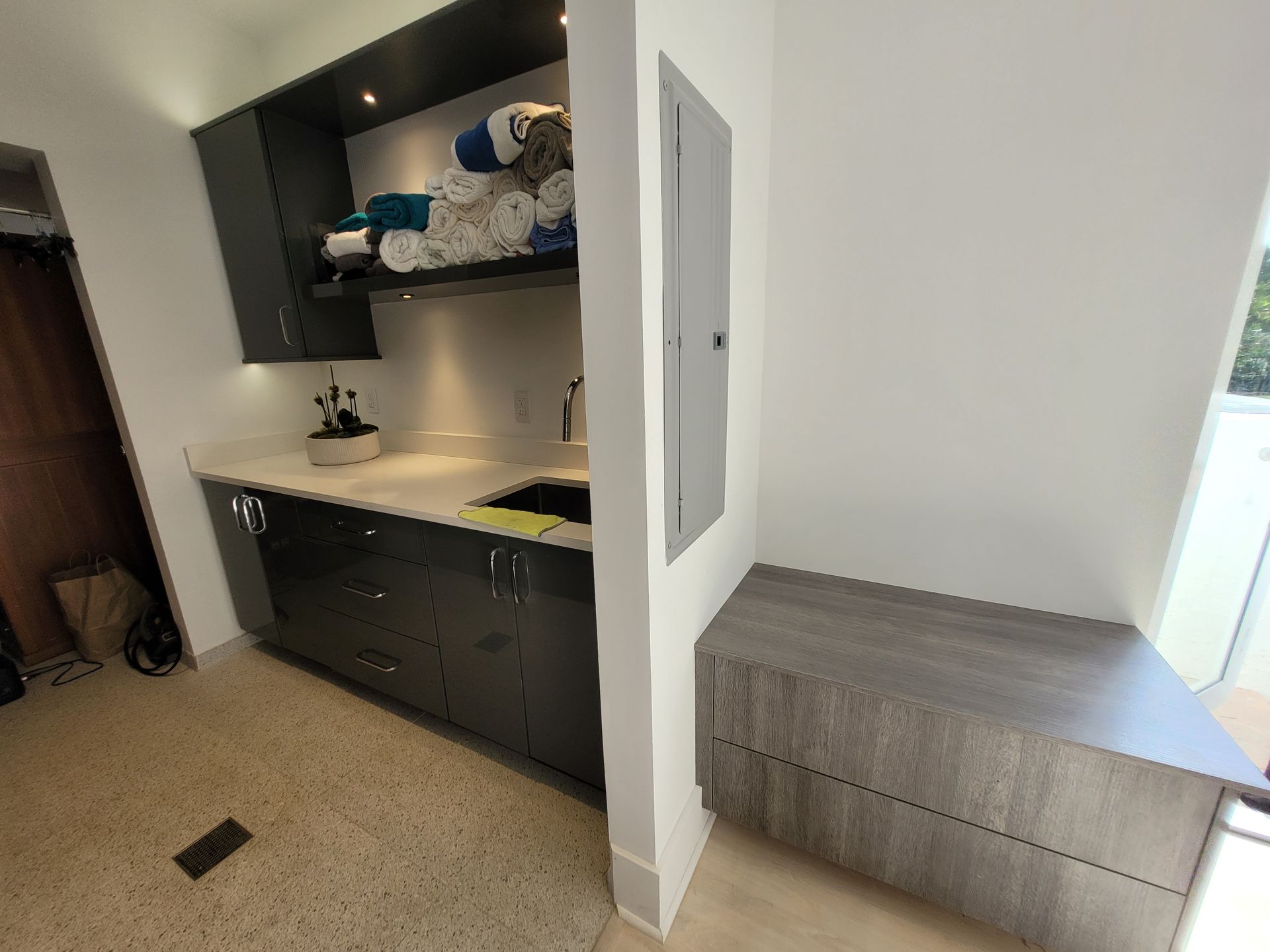 Small utility nook with dark cabinets, sink, shelves, and a gray bench by a bright window.