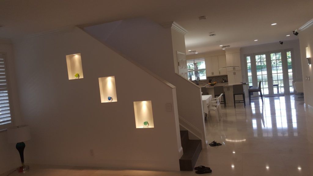 Modern living room with white walls, built-in lit niches, glossy floors, staircase, and open view to a bright entryway