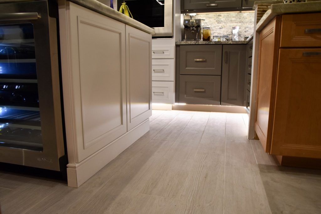 Kitchen aisle between white and wood cabinets, with stainless appliances and a lit sink area in back.