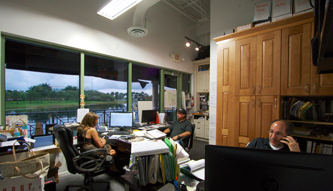 Office with people working at desks, large windows overlooking water, and a wooden cabinet.