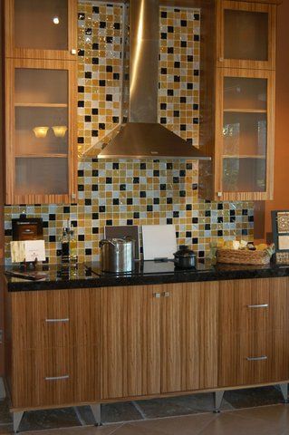 Kitchen cabinets with mosaic tile backsplash, stainless range hood, and black countertop.