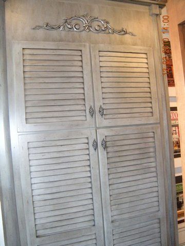 Gray louvered double-door cabinet with decorative carving on top, standing indoors.