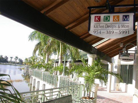 Outdoor walkway under a wooden roof with potted palms and a sign for “Kitchen Designs & More” above the railing