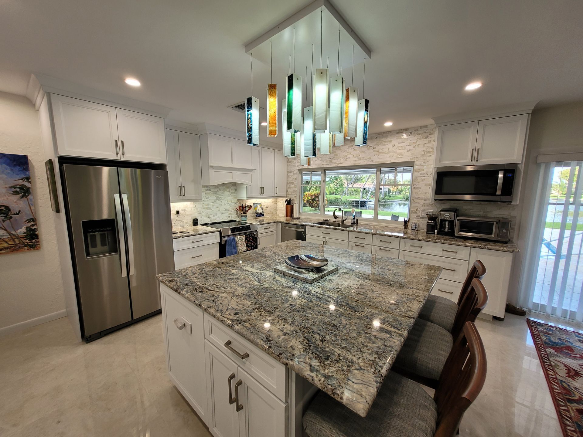 Modern white kitchen with a large granite island, stainless steel appliances, and hanging pendant lights.