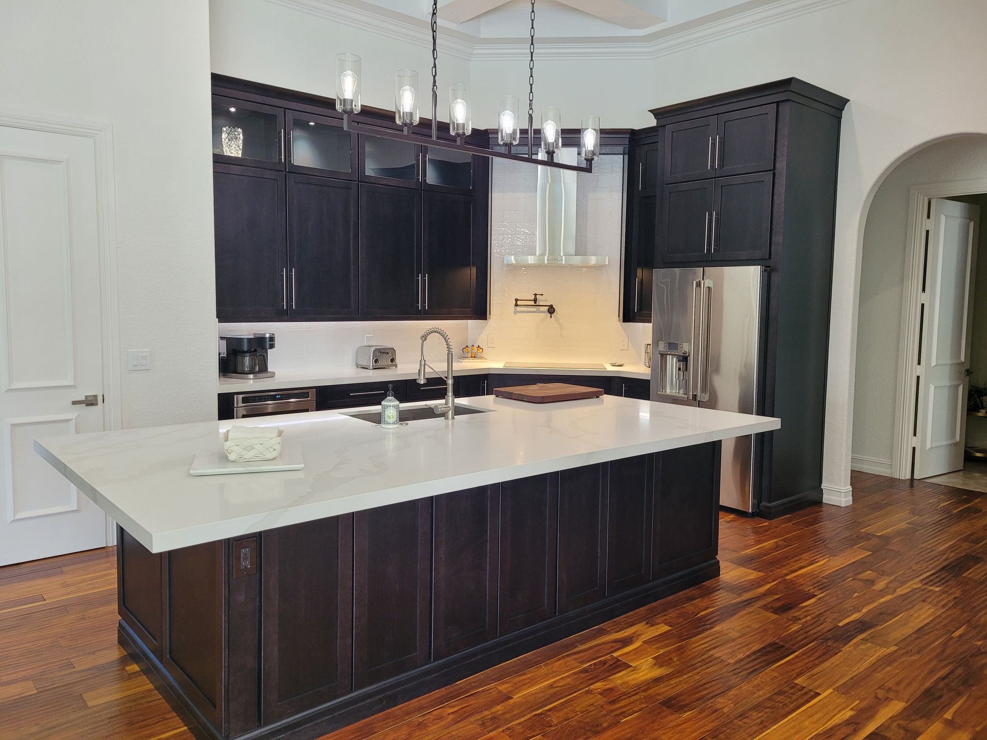 Modern kitchen with dark cabinets, white island, and stainless steel fridge under pendant lights