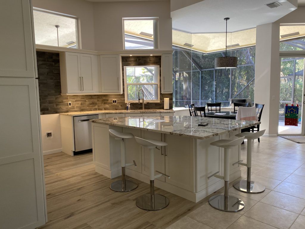 Bright kitchen with white cabinets, island stools, tiled floor, and large windows overlooking trees.
