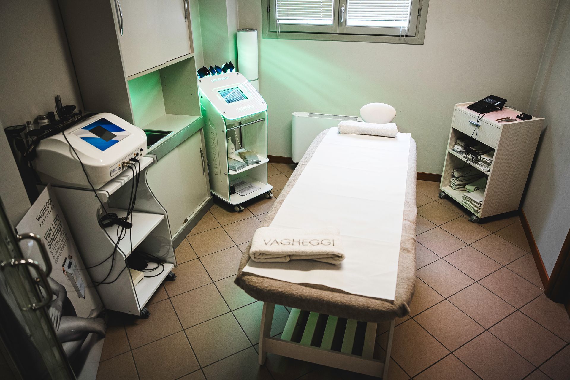Una sala per trattamenti spa professionale con lettino da massaggio, attrezzature per la cura della pelle e un piccolo carrello per i prodotti, su un pavimento piastrellato.