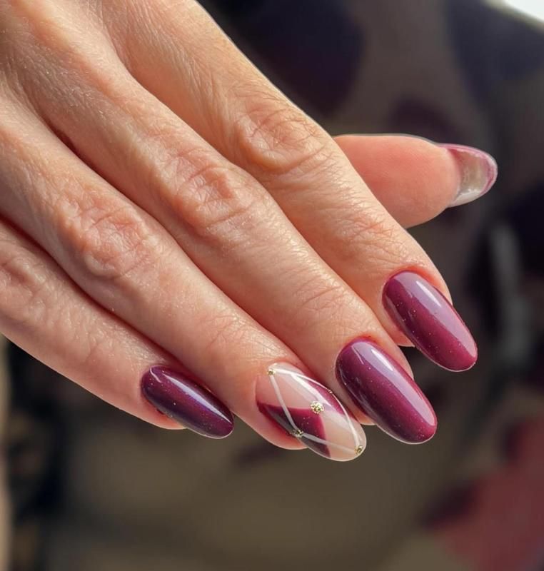 Mano con unghie ovali dipinte di un color prugna scuro, con un'unghia decorata con un disegno geometrico e sottili linee bianche.