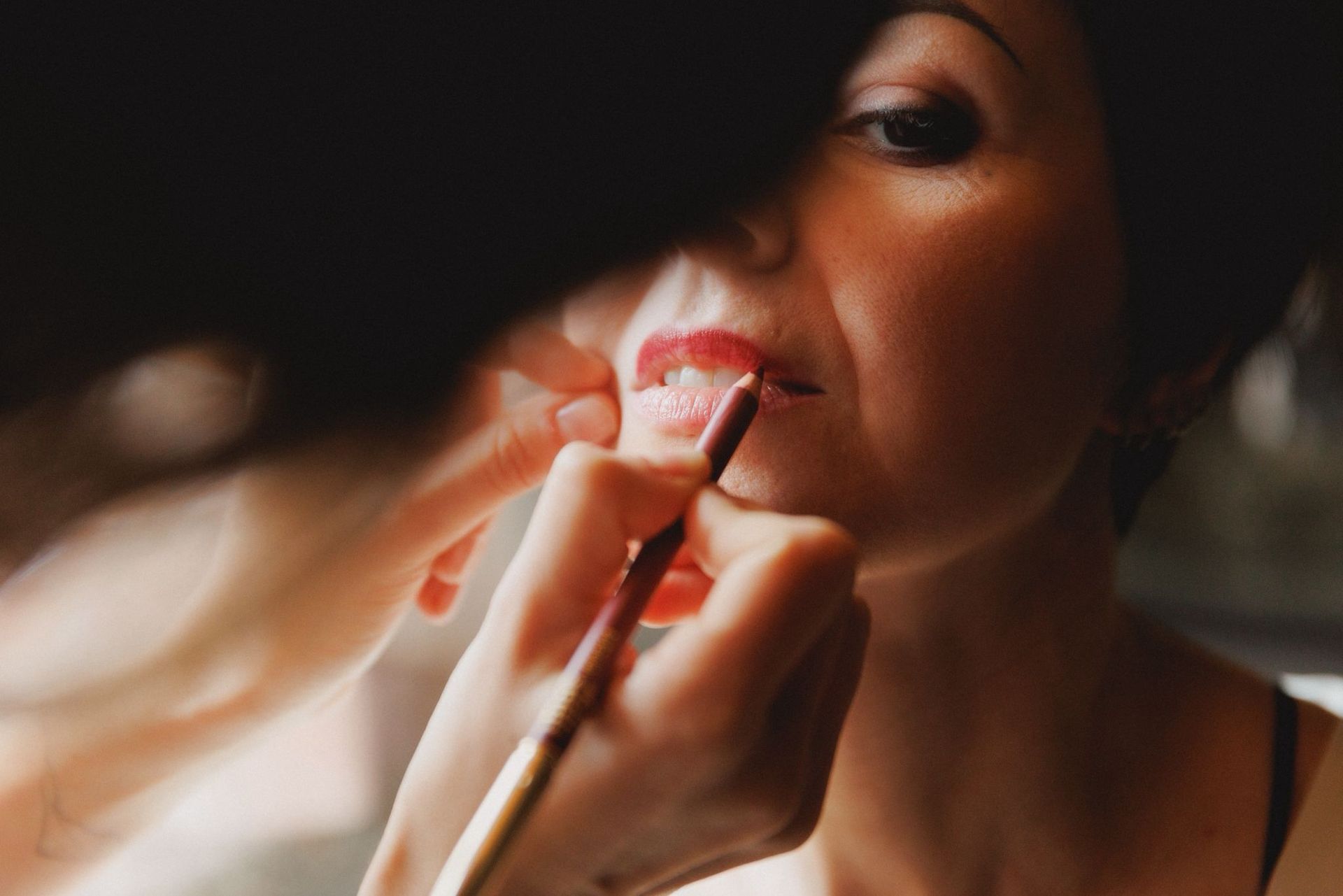 In un primo piano di un servizio fotografico di bellezza, una mano utilizza una matita per labbra scura per delineare il contorno delle labbra di una persona.