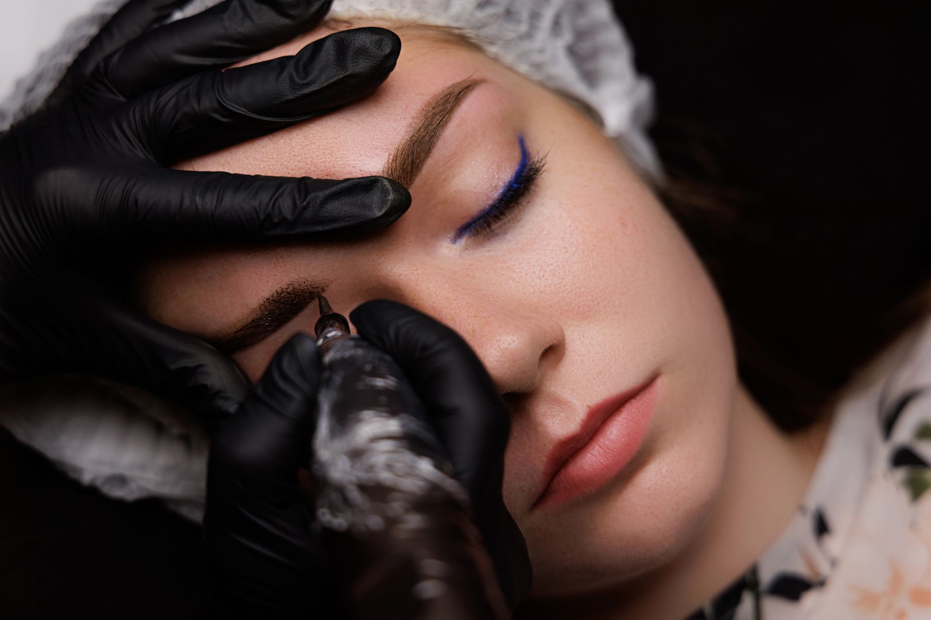 Un professionista con indosso guanti neri utilizza una macchinetta per tatuaggi cosmetici per applicare il pigmento alle sopracciglia di una cliente.