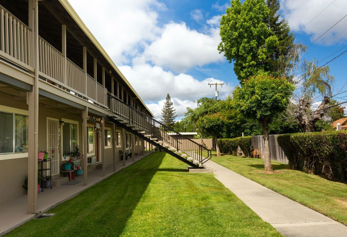 Apartment building with outdoor walkways, stairs, and green lawn under a partly cloudy sky.