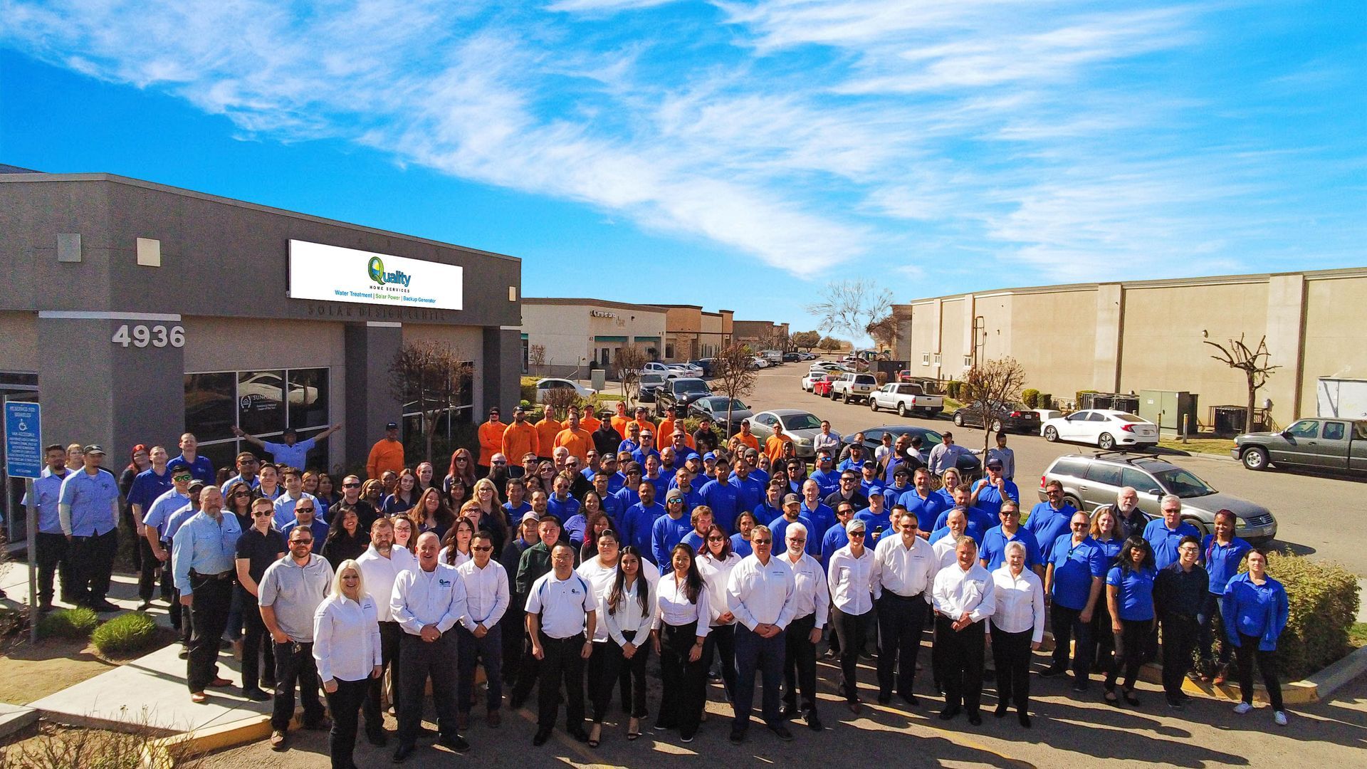 Large group of people in blue, orange, and white shirts in front of a building with the address