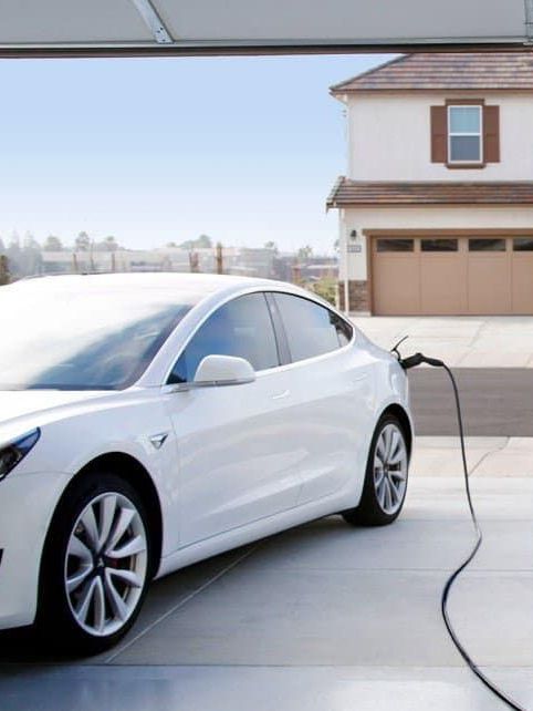 White Tesla electric car charging in a garage driveway in front of a house.