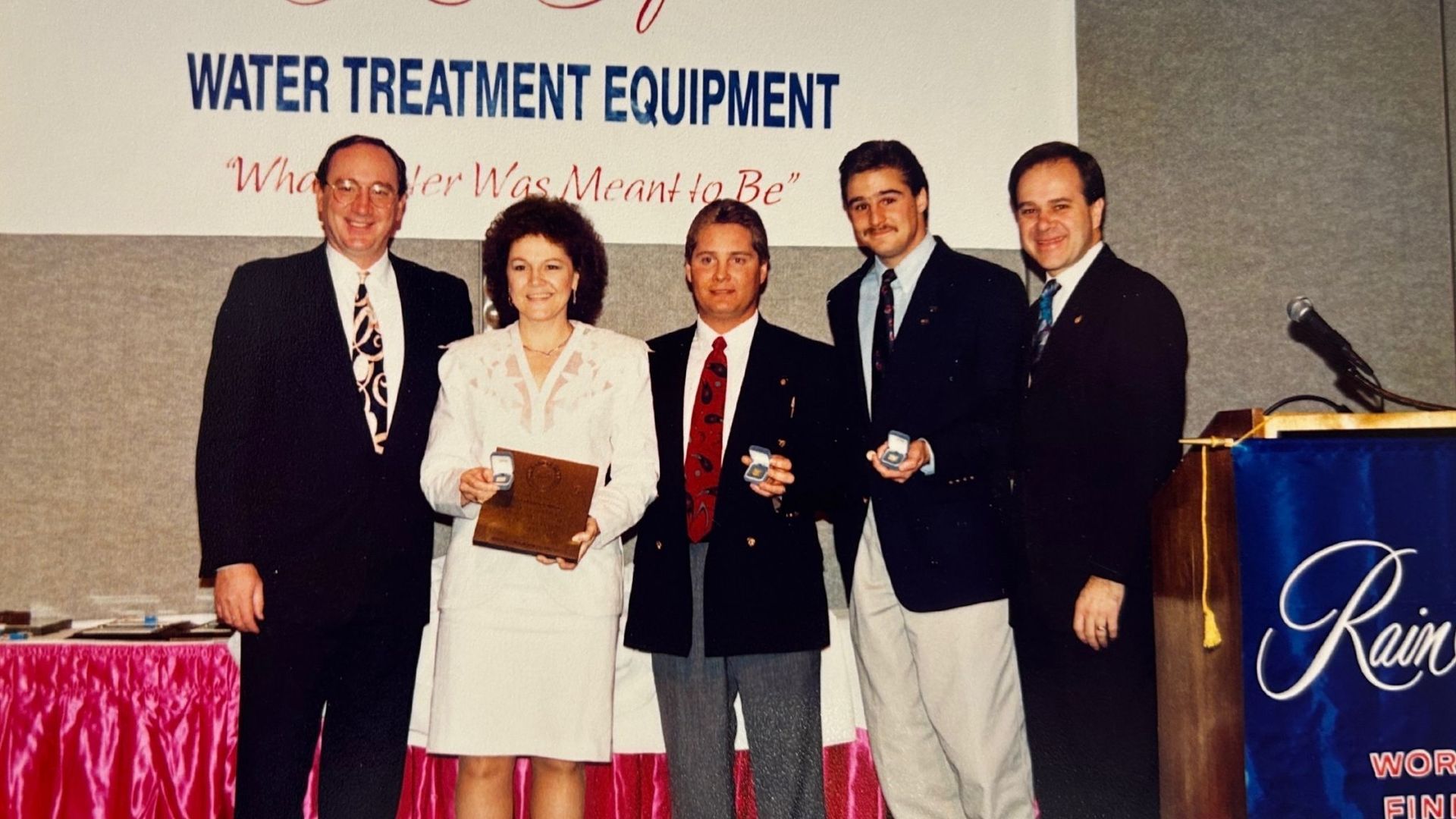 Five people in suits stand at an event, holding awards, with a banner that reads