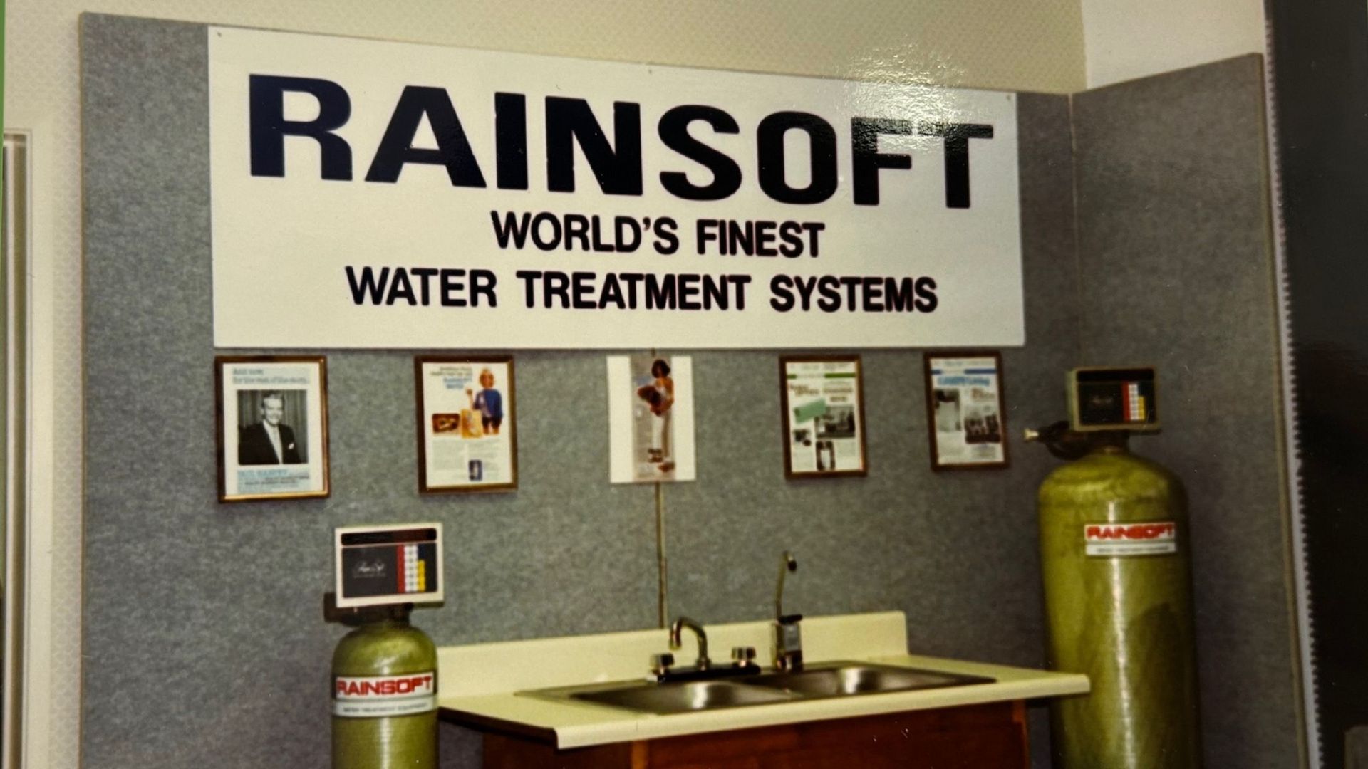 Rainsoft water treatment systems display: sign, brochures, sink, and water tanks.