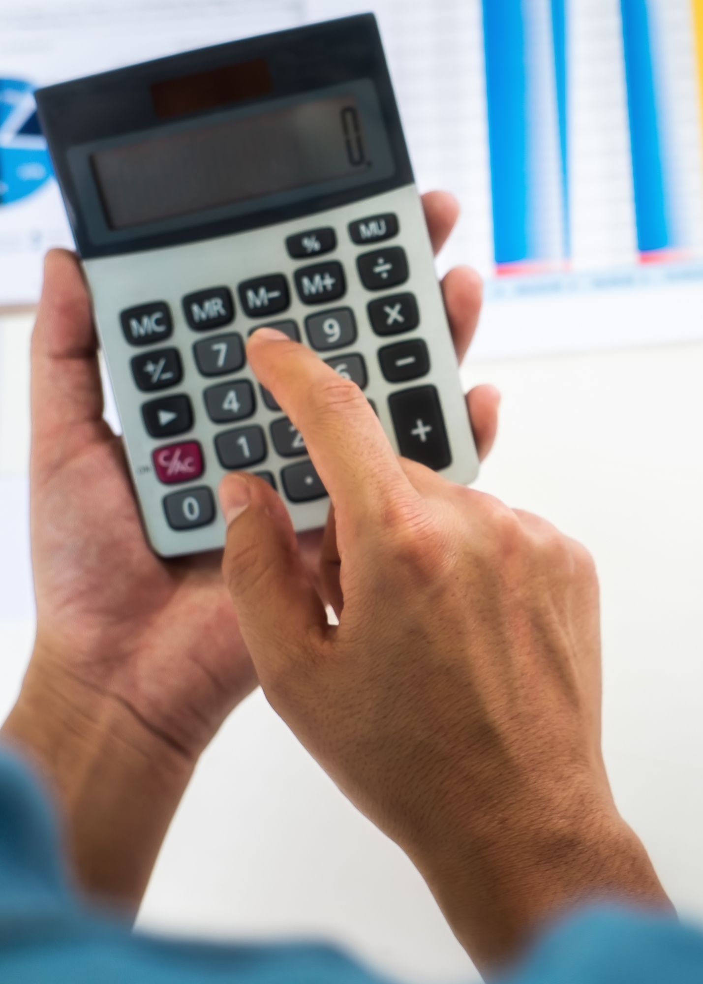 Person's hands holding a calculator, finger pressing the number 7. Charts visible in the background.
