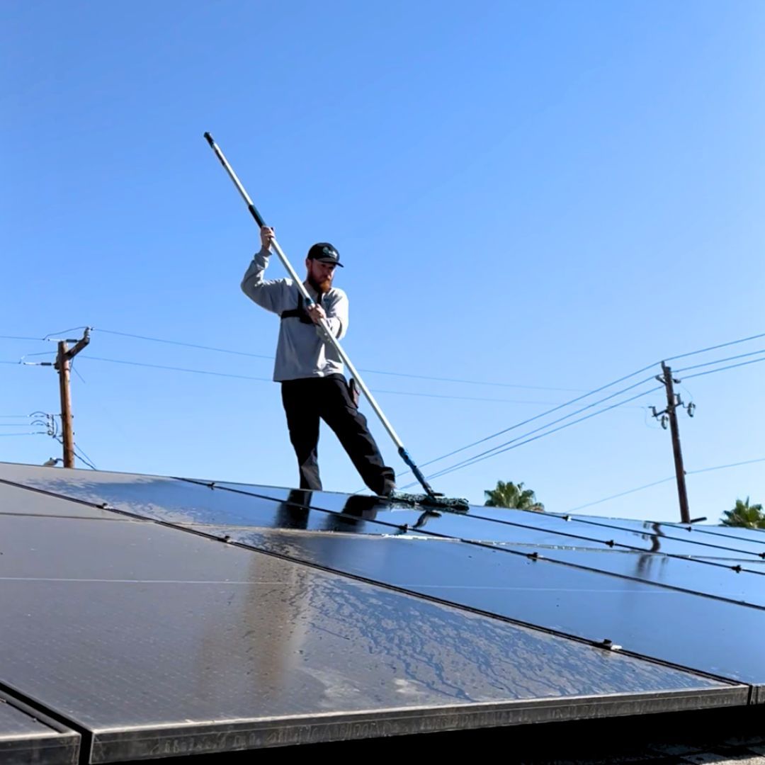 Person cleans solar panels on a rooftop with a long-handled brush under a blue sky.