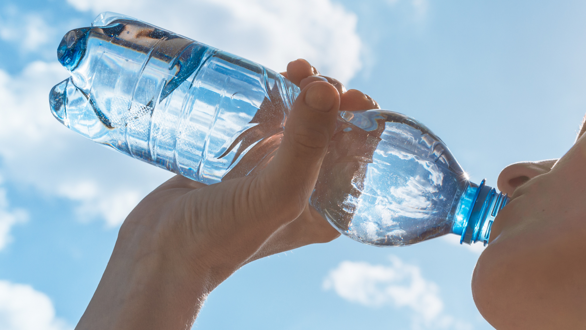 Person drinking water from a clear plastic bottle against a blue sky.