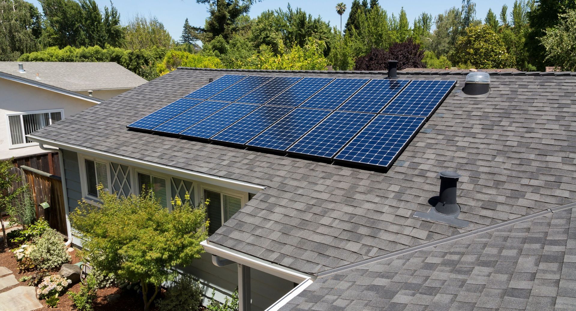 Rooftop solar panels on a gray shingle house surrounded by trees under a clear sky