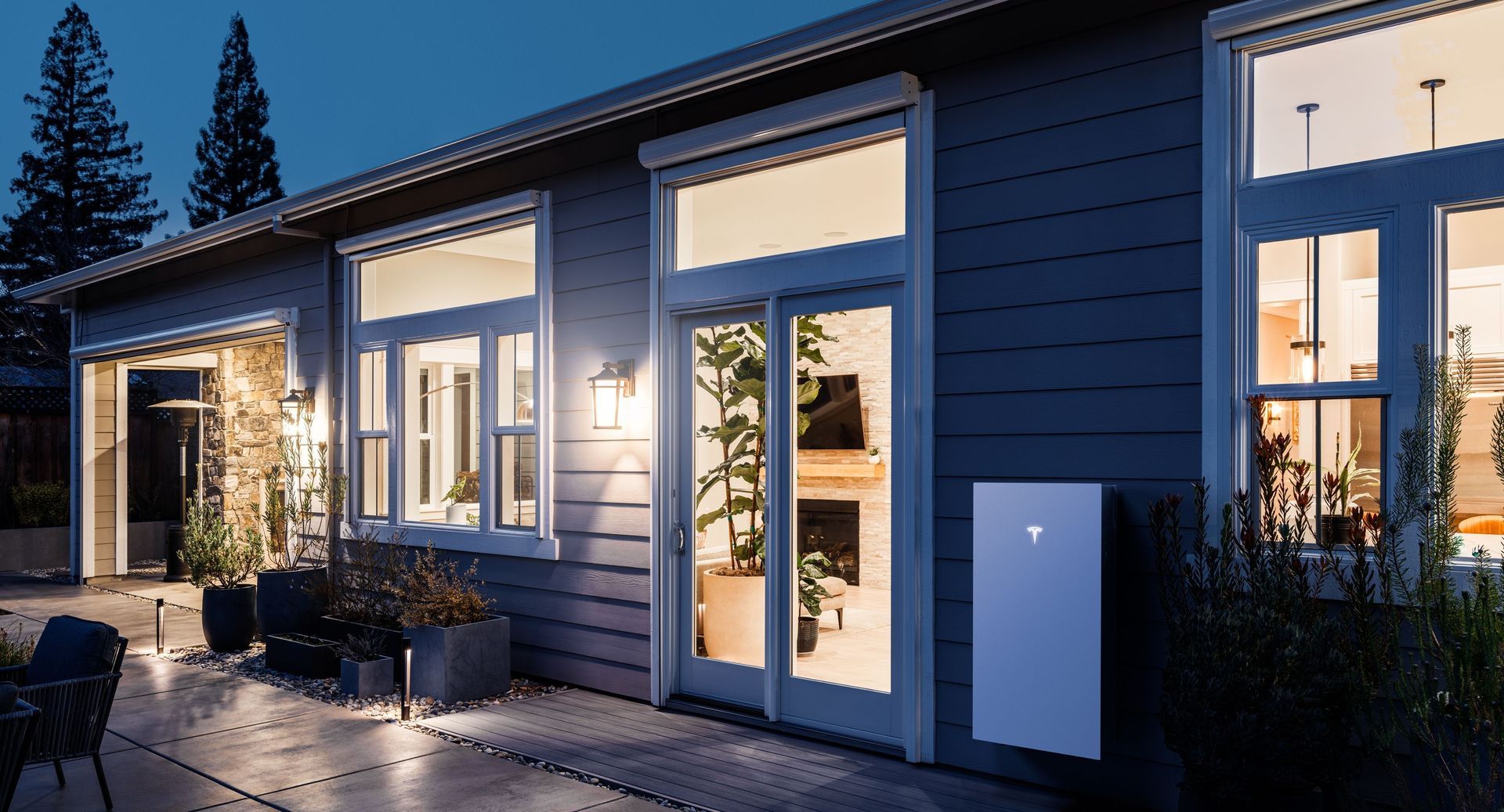 Modern blue house exterior at dusk with lit windows and a glass door leading to a patio