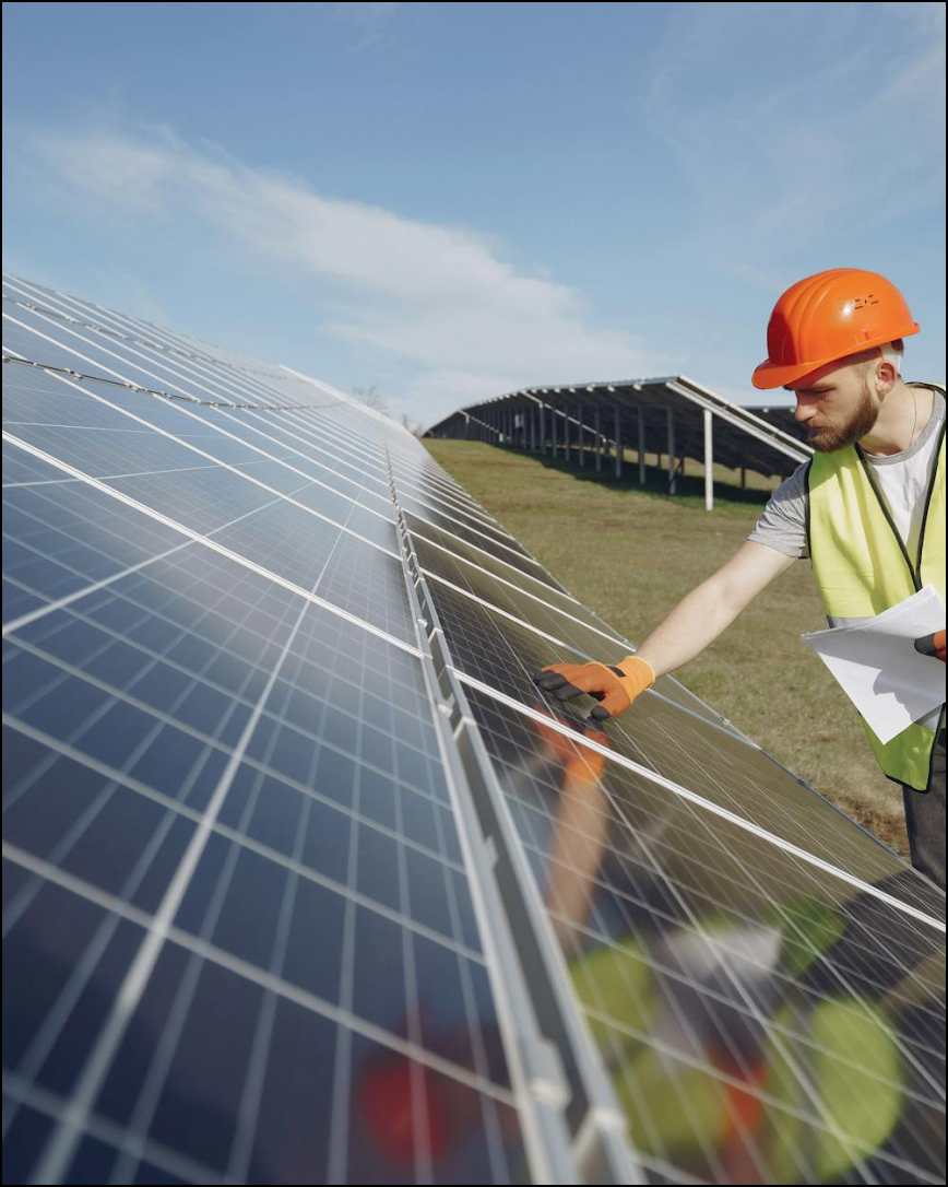 A worker in orange hard hat and safety vest inspects solar panels in a field.