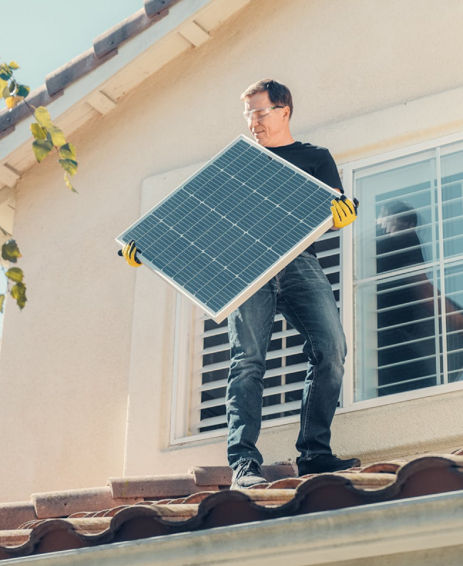 Man on a rooftop holding a solar panel. He wears safety glasses, yellow gloves, and jeans. Sunlight.