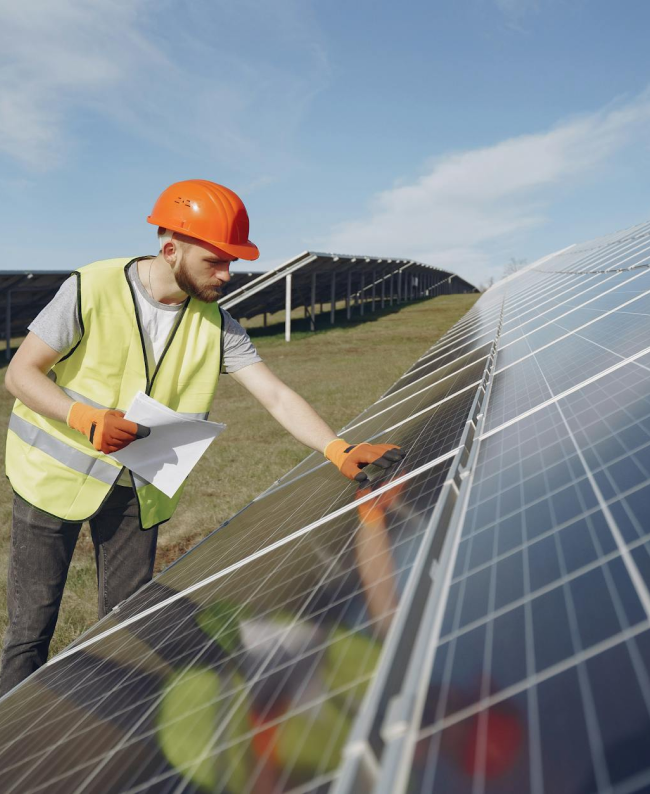 Solar panel technician inspecting solar panels outdoors, wearing orange hard hat and safety vest.