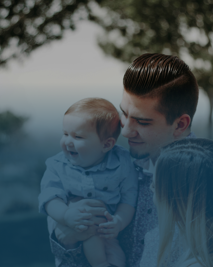 Man holding a baby, looking at the child. Woman's shoulder and hair visible. Blurred outdoor setting.