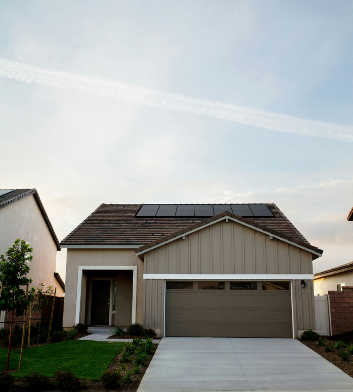Tan house with brown roof, solar panels, and concrete driveway against a pale blue sky.