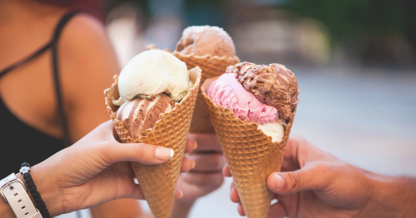 Three hands holding waffle cones with scoops of vanilla, pink, and chocolate ice cream outdoors