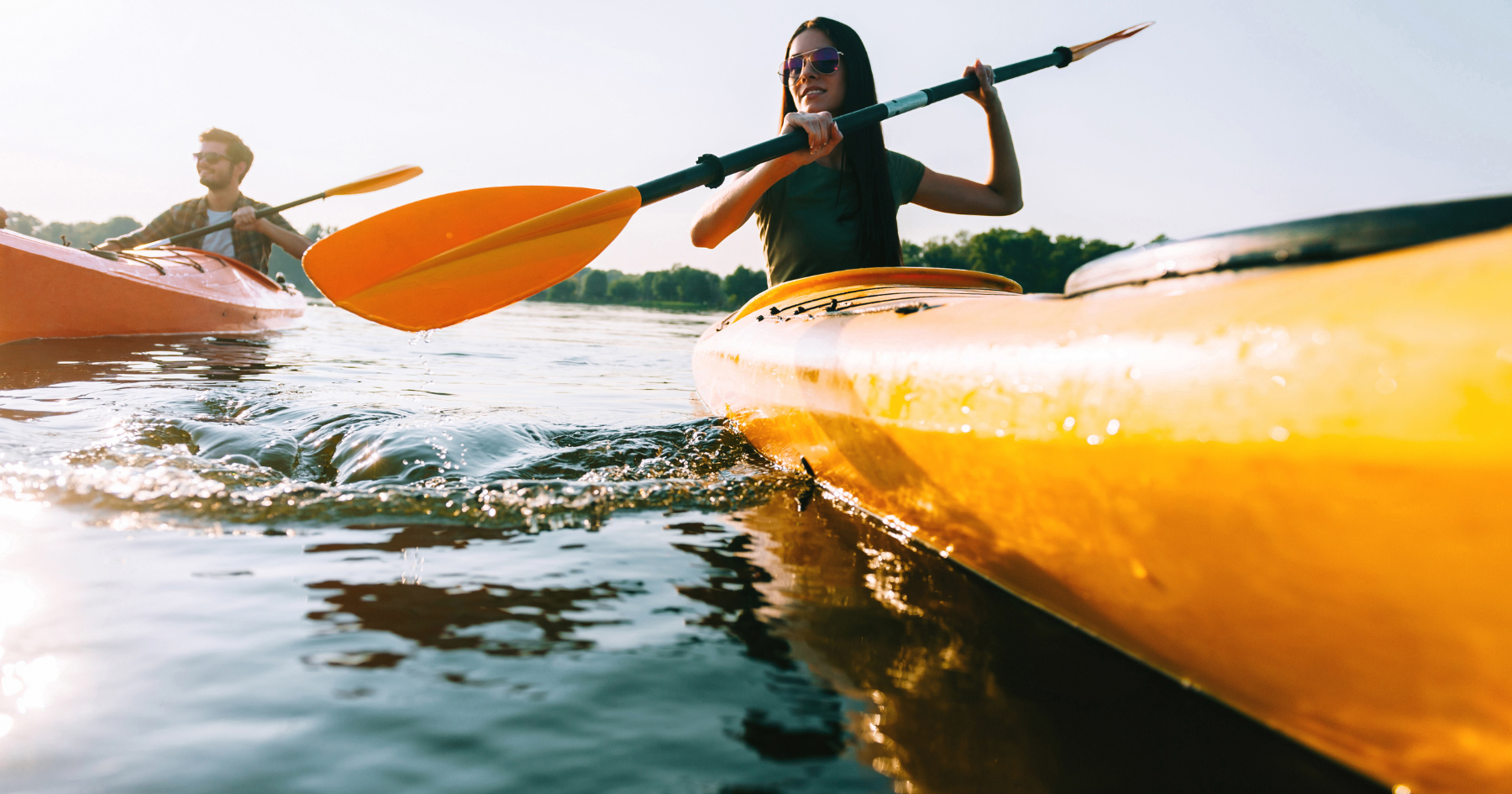 Two people kayaking on calm water, one in a bright yellow kayak with orange paddles at sunset