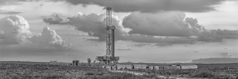 A black and white photo of an oil rig with puffy clouds in the sky.