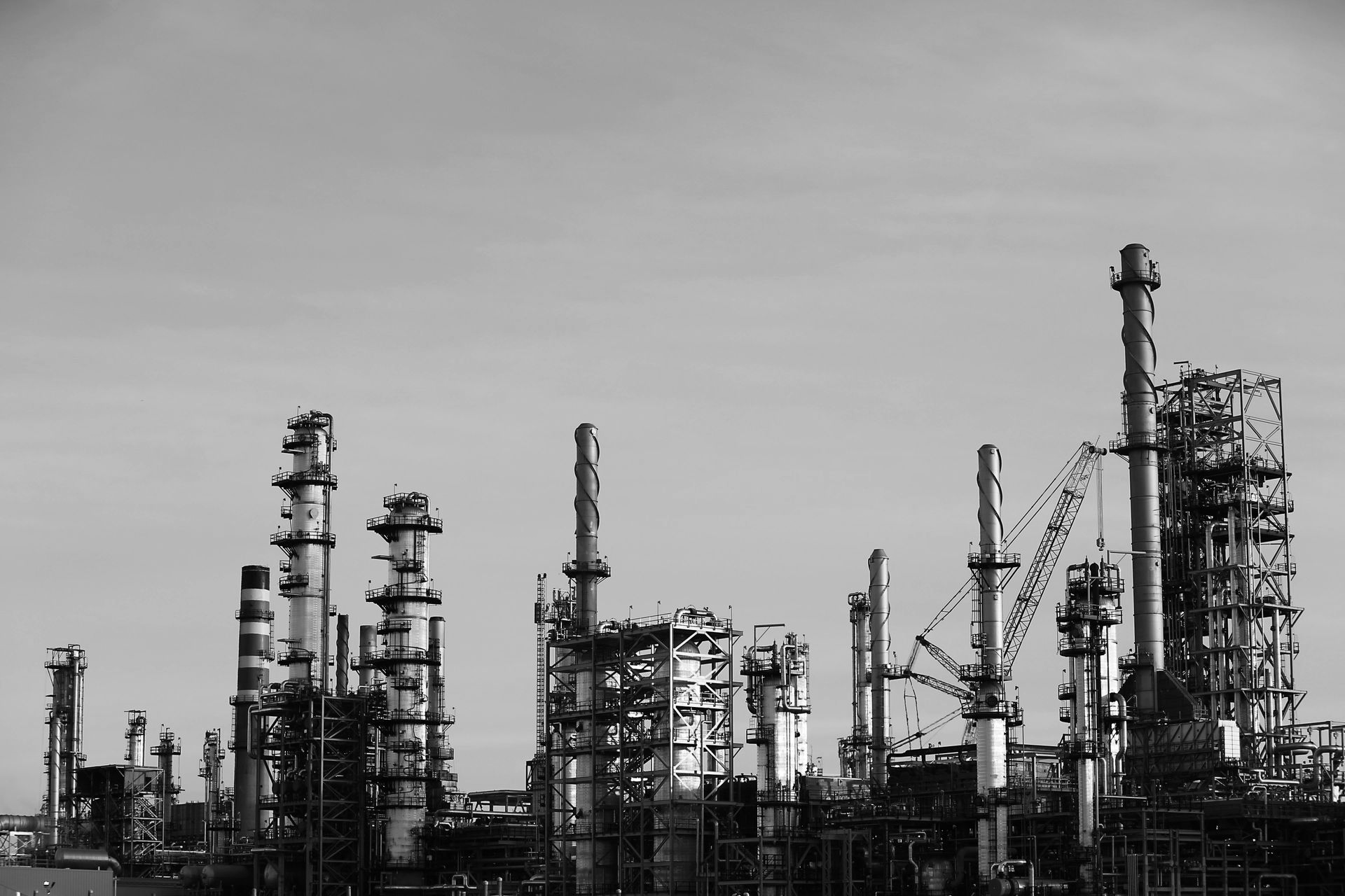 Industrial oil refinery complex against a pale blue sky. Steel structures, smokestacks, and piping are visible.