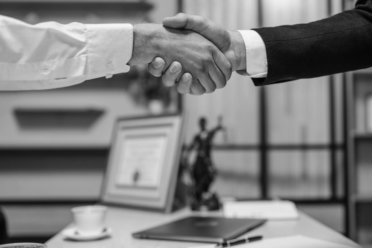 Two people shaking hands over a desk, likely sealing a deal, in an office setting.