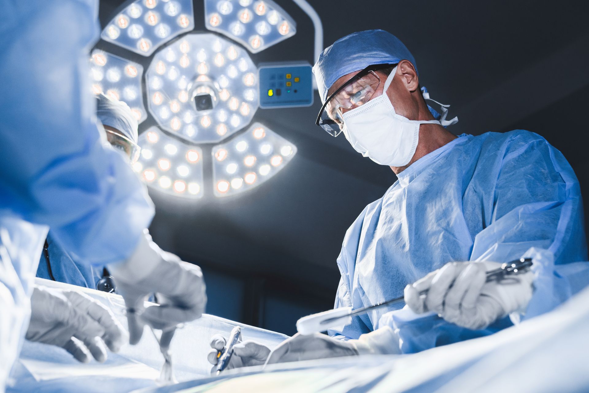 Surgeon in blue scrubs, mask, and goggles operates under bright surgical lights.