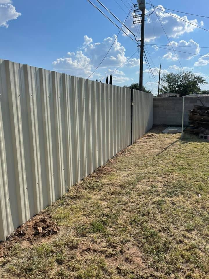 White corrugated metal fence beside a grassy dirt strip under a blue sky with clouds