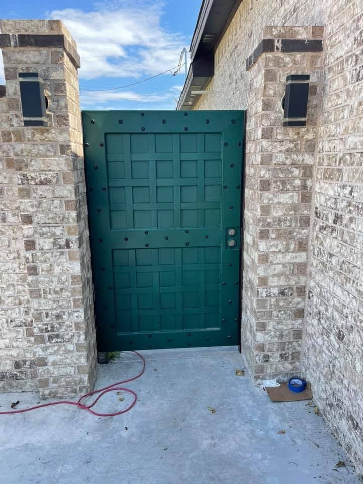 Green metal gate between two stone pillars, with a concrete driveway and coiled red hose on the ground.