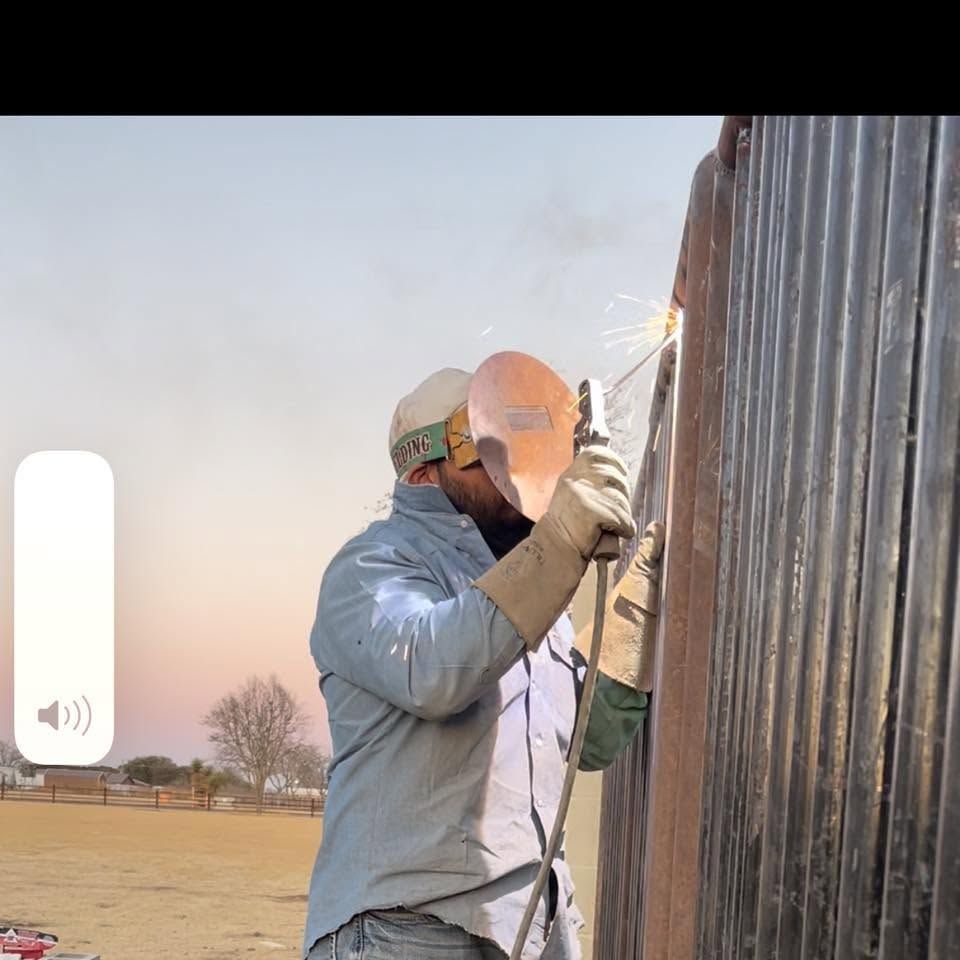 Worker welding a corrugated metal fence outdoors, sparks flying at dusk.