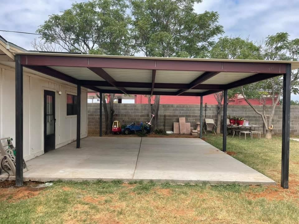 Covered concrete carport beside a white house with black posts and trees in the yard