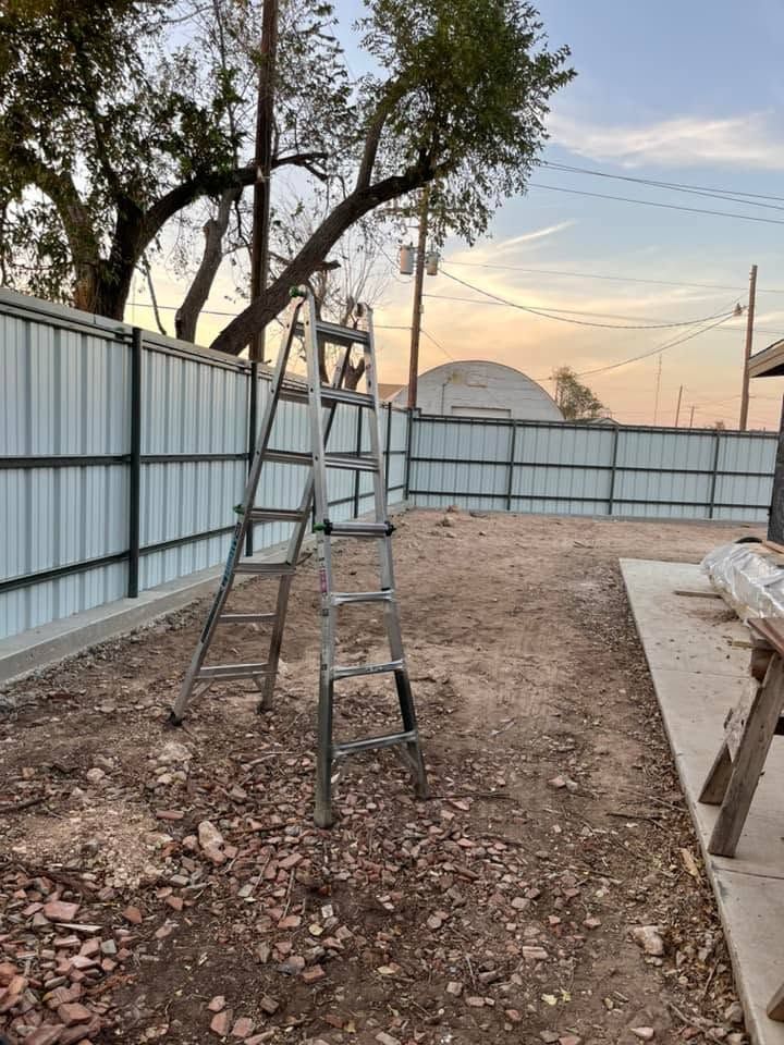 Ladder leaning against a fence in a dirt yard at sunset, with trees and a metal wall in the background