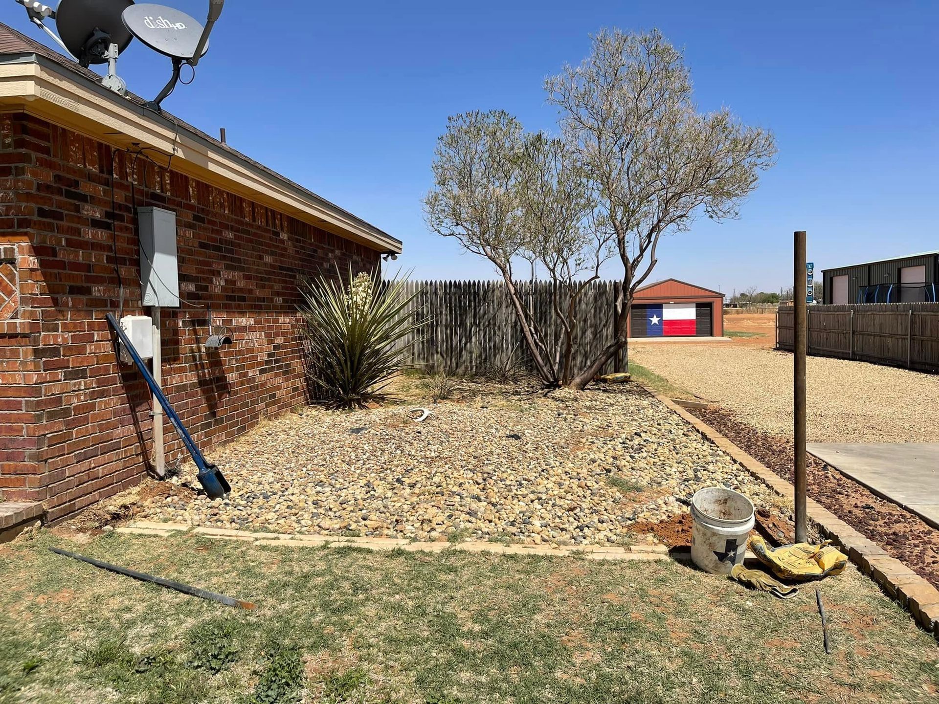 Backyard beside a brick house with a tree, gravel bed, lawn, and a red shed in the distance
