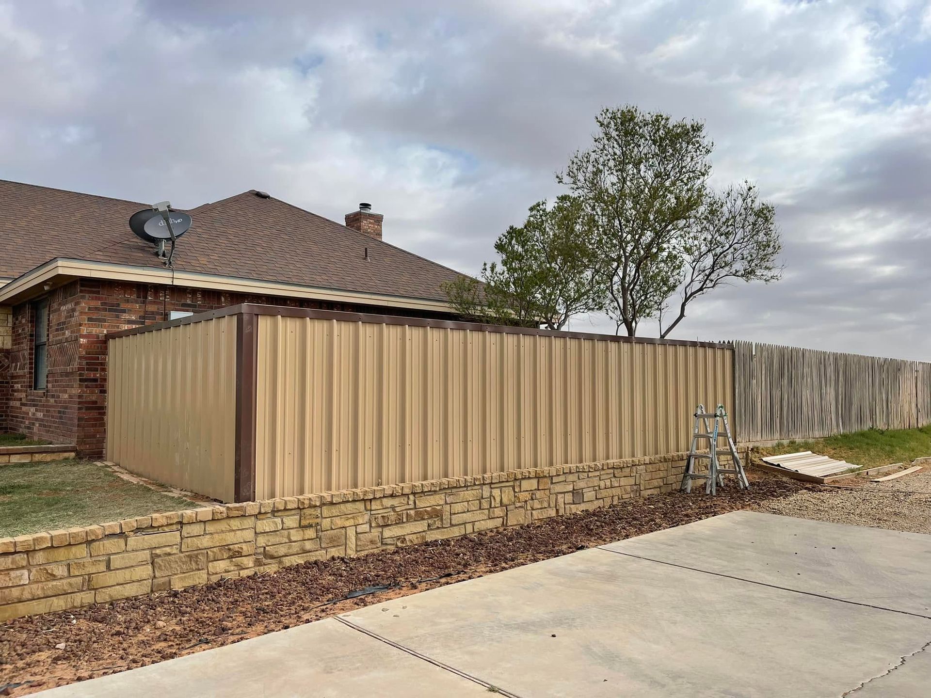 Tan corrugated metal fence and shed beside a brick house on a suburban street under cloudy skies
