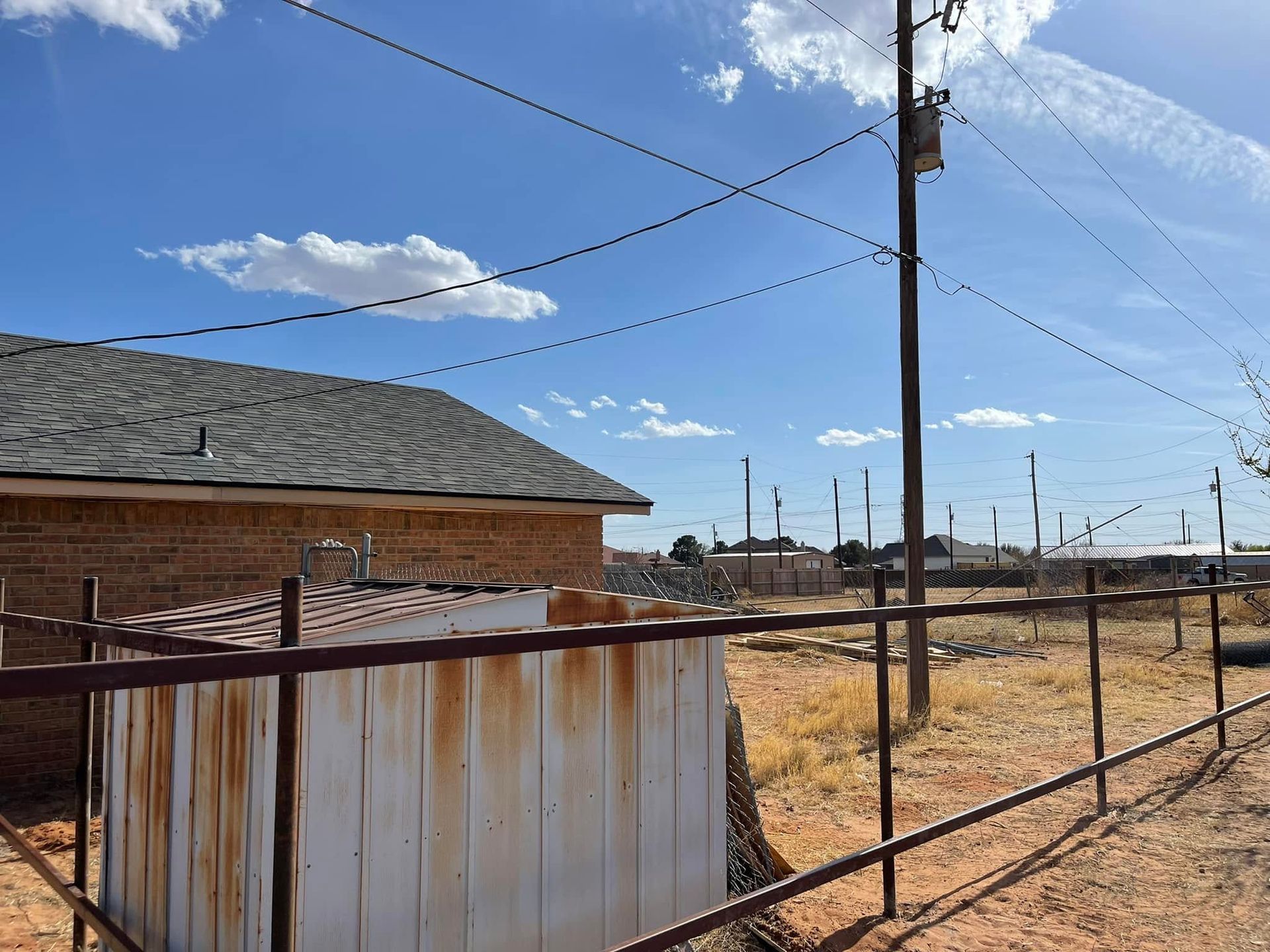 Rustic yard with a corrugated metal fence, brick building, utility poles, and dry grass under a blue sky.