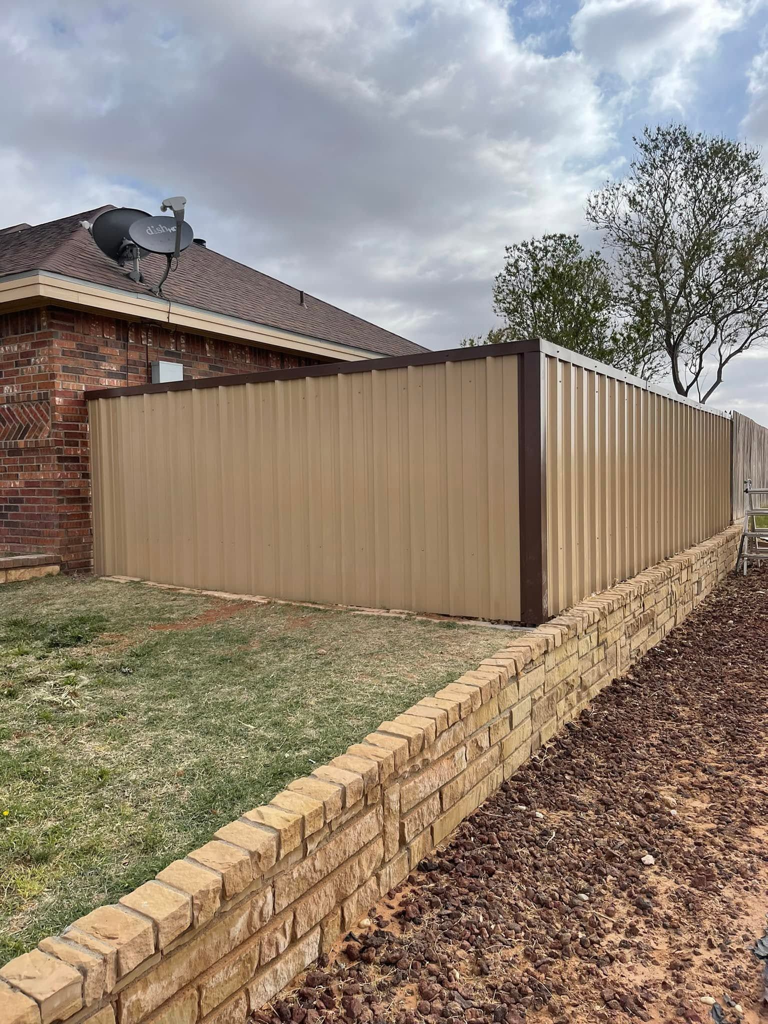 Tan metal fence beside a brick house along a sloped yard under cloudy sky