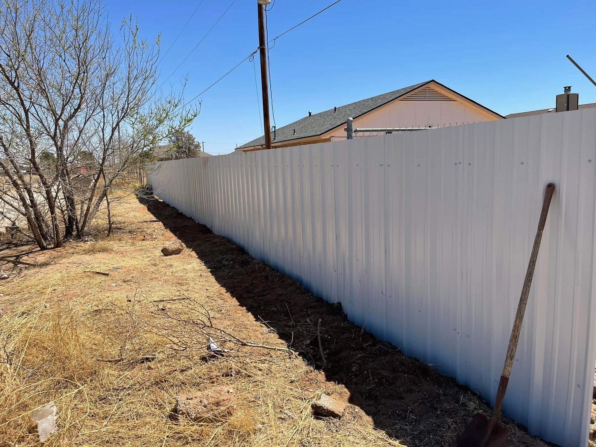 White fence along a dry dirt yard beside a house under a clear blue sky